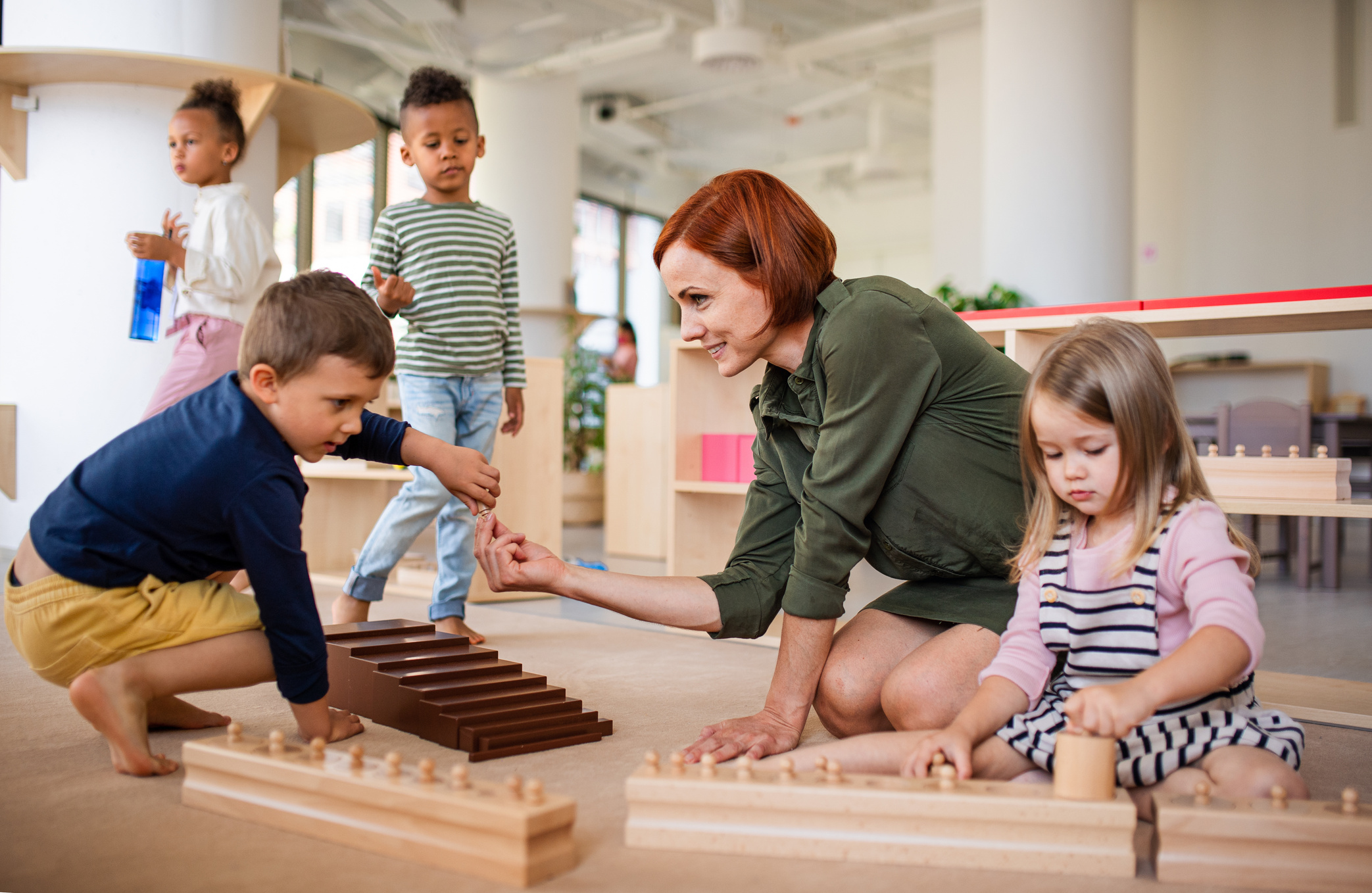 Group of Small Nursery School Children with Teacher Sitting on Floor Indoors in Classroom, Montessori Learning.